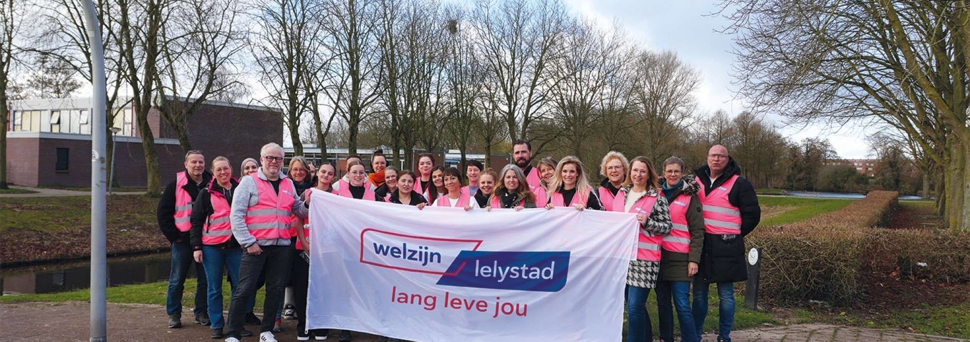 Mensen met roze hesjes houden vlag met logo van Welzijn Lelystad vast.