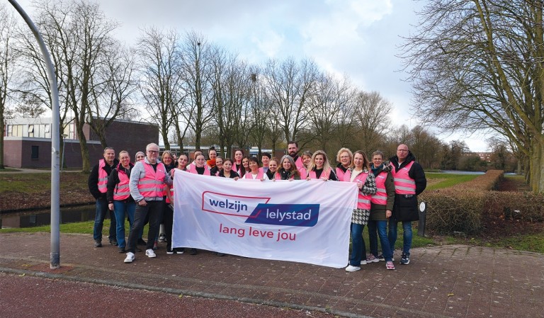 Mensen met roze hesjes houden vlag met logo van Welzijn Lelystad vast.