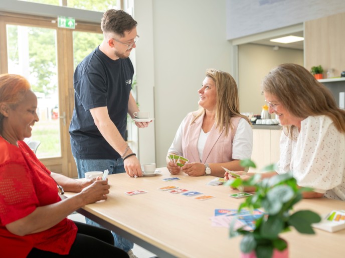 Drie vrouwen doen een spelletje aan tafel, een man serveert koffie.