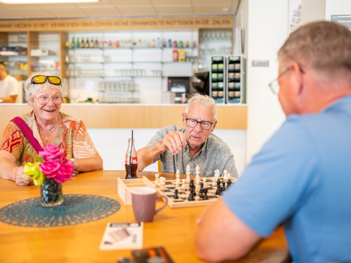Twee mensen spelen potje schaak en een vrouw kijkt mee.