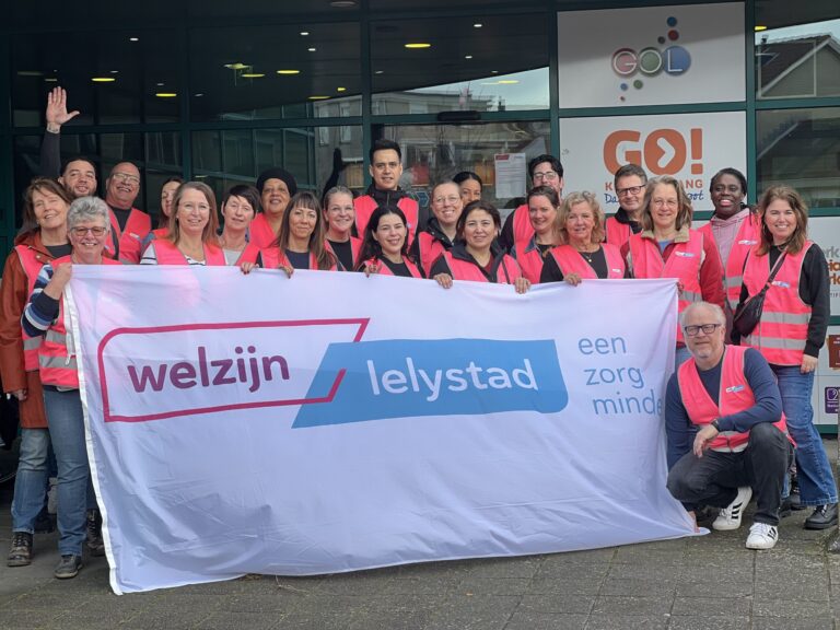 Mensen in roze hesjes houden een vlag van Welzijn Lelystad vast.